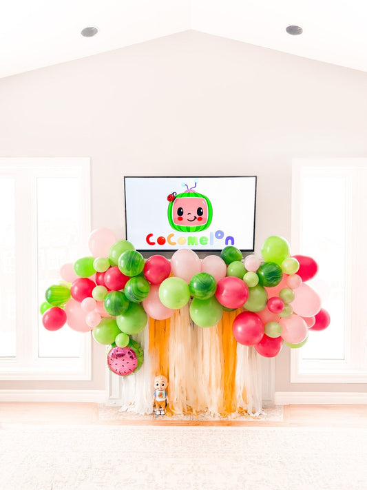 Pink, lime green, and peach balloon garland arch with a fruit face sign at center