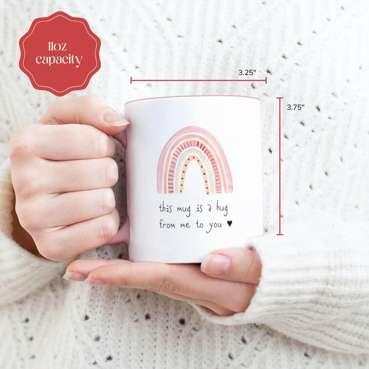 Hands holding a small white ceramic mug with pink handle and rainbow hug quote
