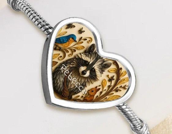 Heart charm bracelet close-up with silver snake chain and raccoon artwork; name Rebecca