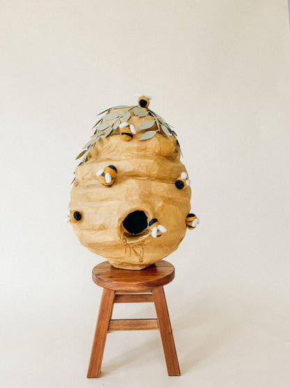 Beehive piñata on wooden stool, leaf crown and felt bees on tan paper surface