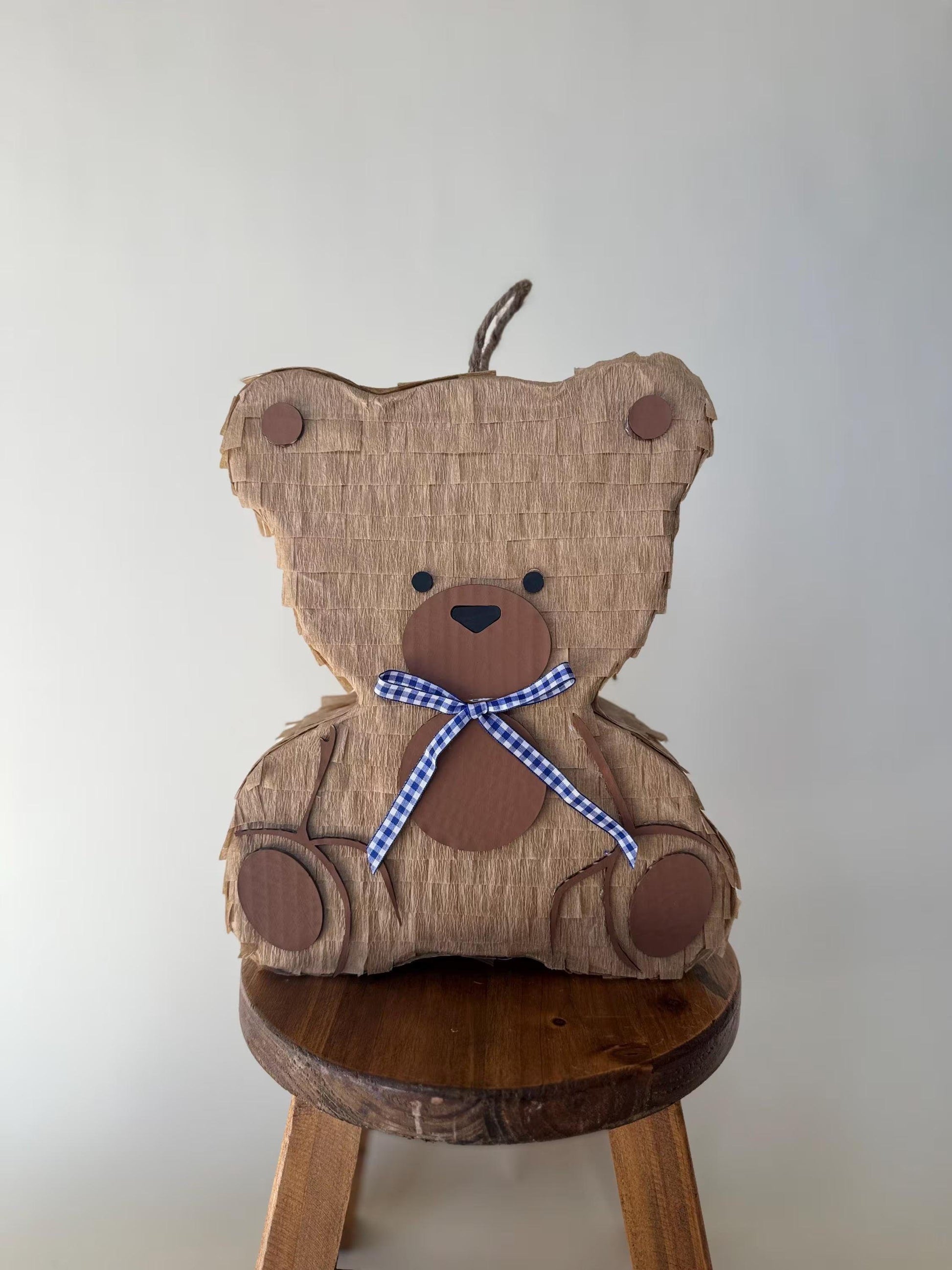 Teddy bear piñata front on round stool, tan cardboard, blue plaid bow, paw details