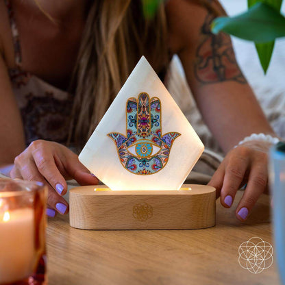 Hands placing a glowing pyramid mood lamp with Hamsa panel on wooden base