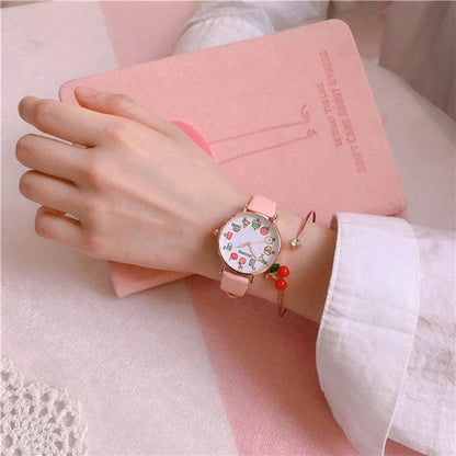 Pink strap wristwatch with food-themed dial and cherry charm bracelet on wrist