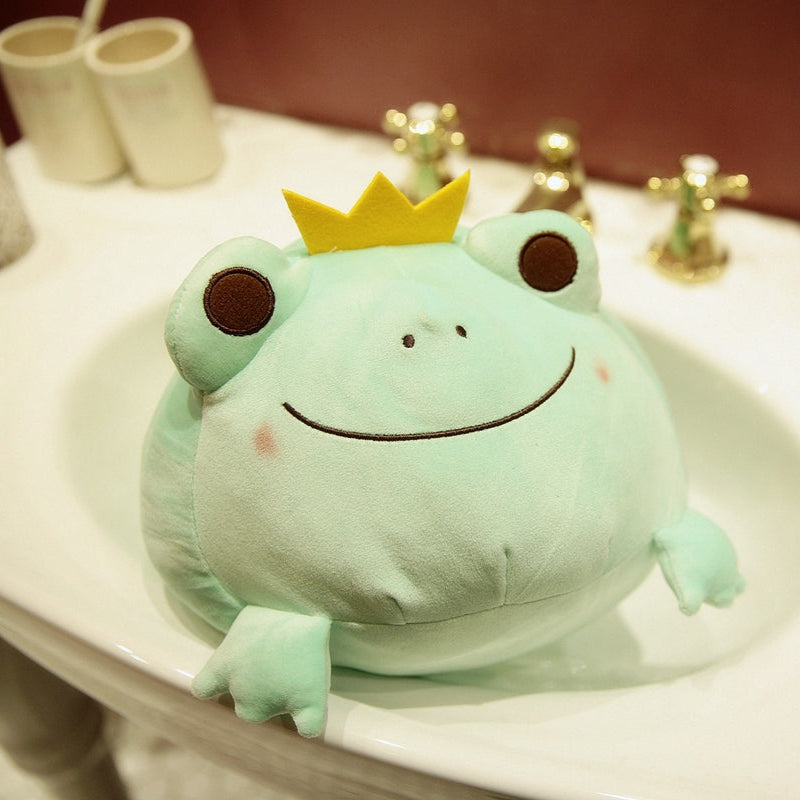 Green frog plush with yellow crown and smiling embroidered face, lying on table